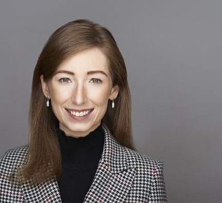 Dr. Christine A. Ryan smiles at the camera against a gray background. She has shoulder-length reddish-brown hair, white skin, green eyes and wears a black turtleneck under a houndstooth coat.